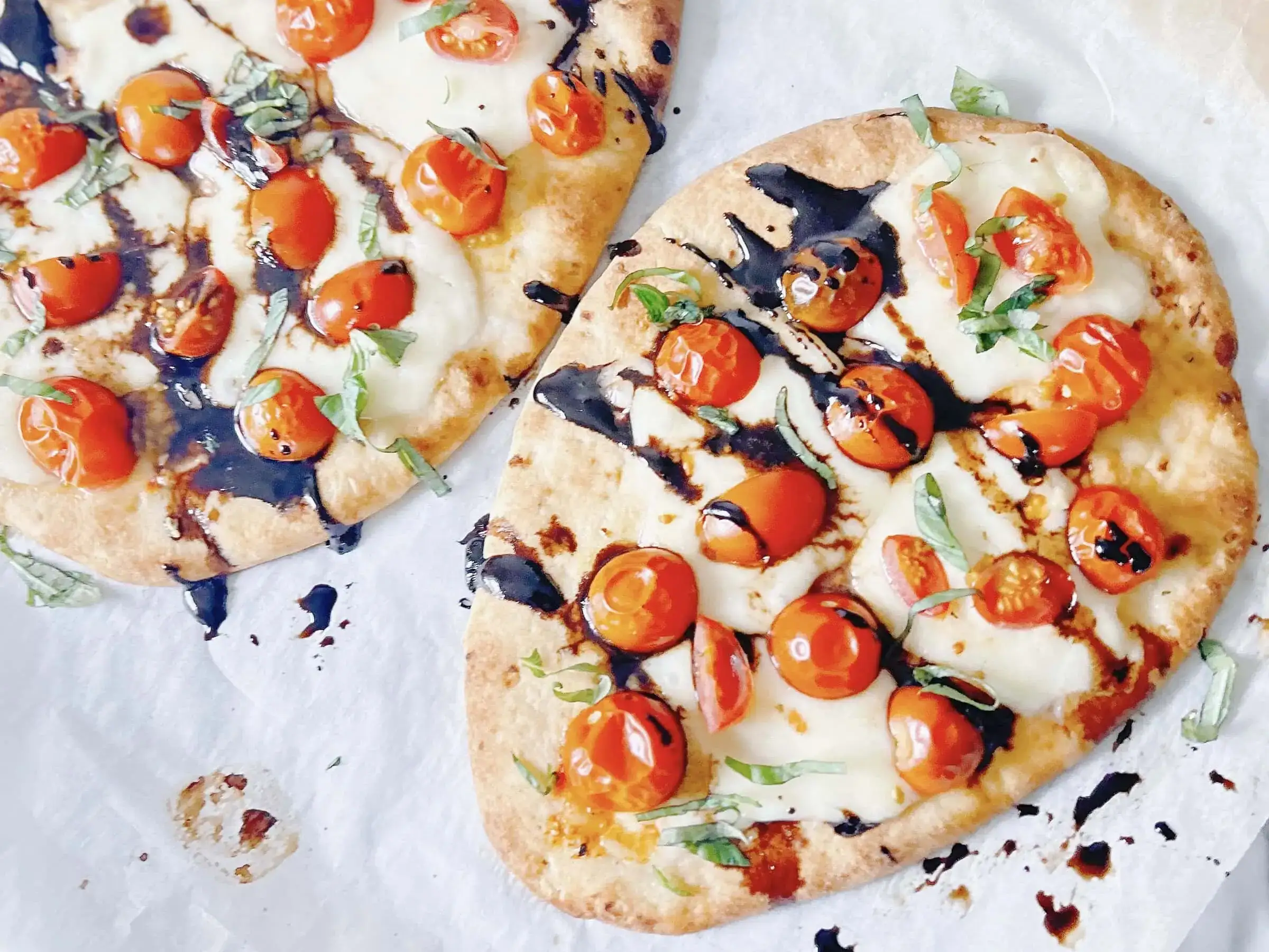 Two flatbreads topped with melted cheese, cherry tomatoes, fresh basil, and a drizzle of balsamic glaze on parchment paper.