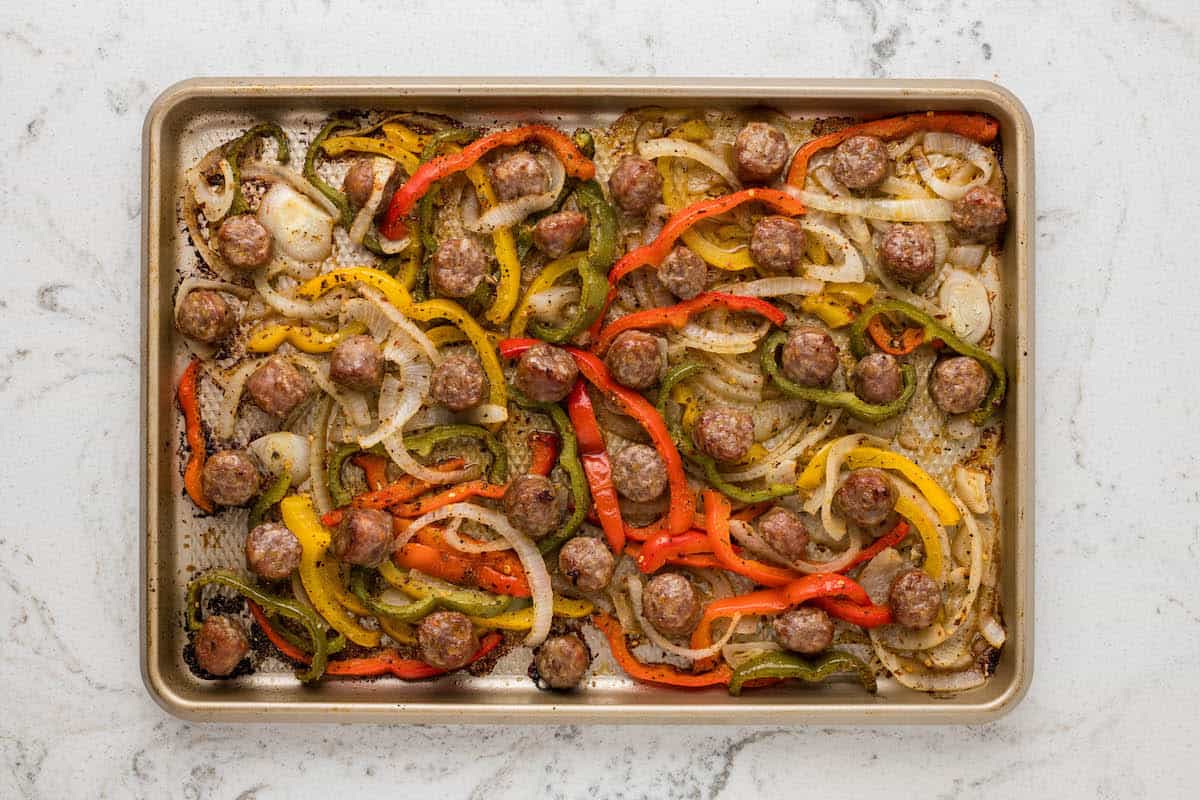 Baked sausage pieces with sliced red, yellow, and green bell peppers and onions spread on a metal sheet pan.