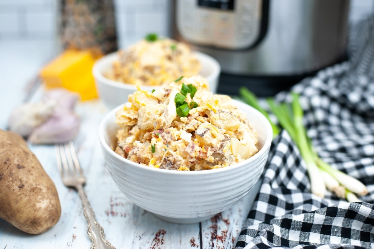 A white bowl filled with potato salad garnished with green onions, with another bowl, potatoes, utensils, and an instant pot in the background.