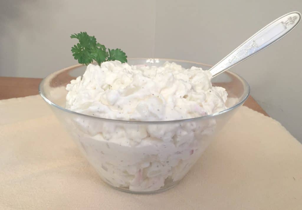 A glass bowl filled with creamy potato salad, garnished with parsley and served with a silver spoon, sits on a light-colored surface.