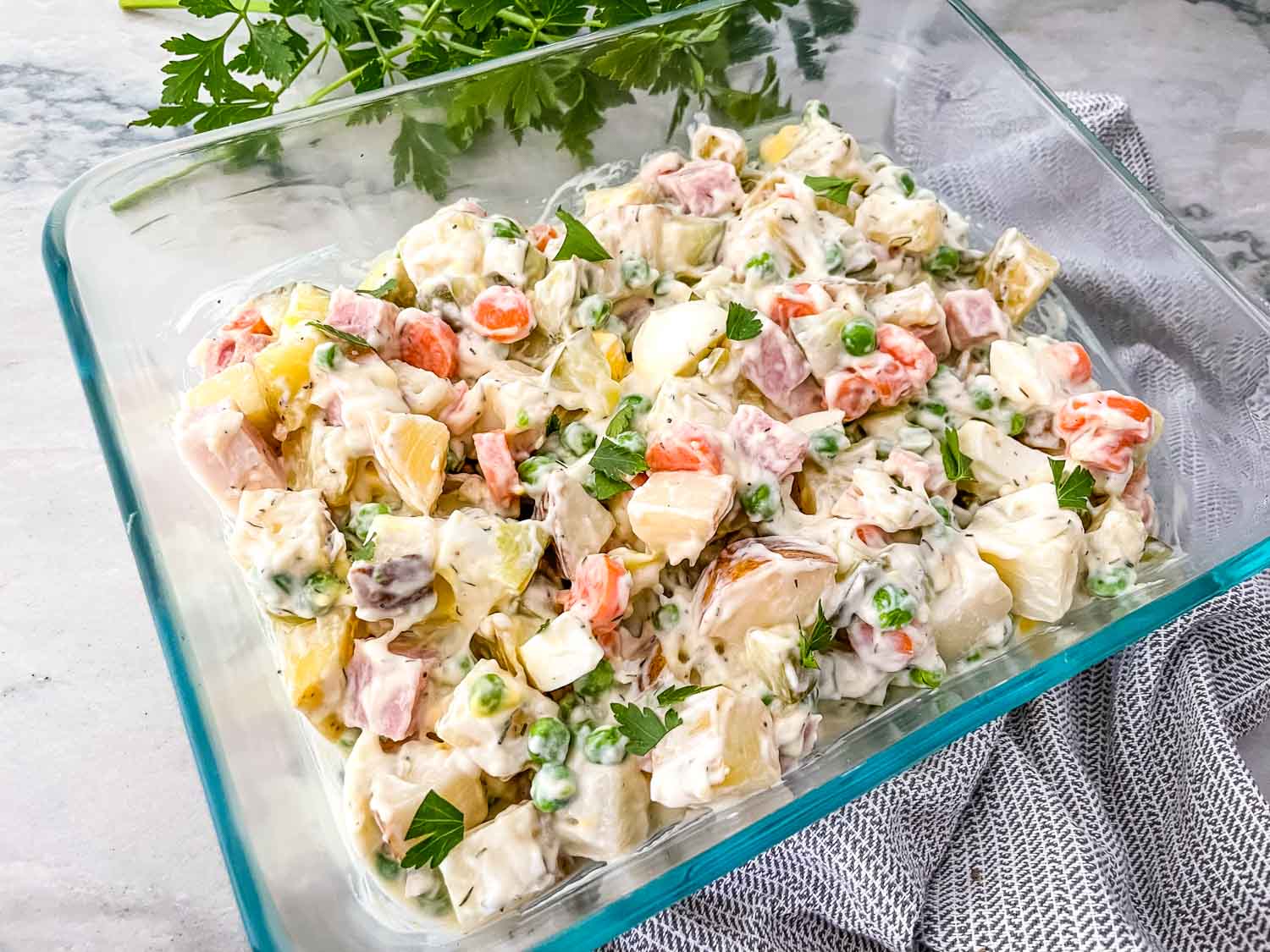 A glass dish filled with creamy salad containing diced vegetables, ham, peas, and garnished with fresh parsley.