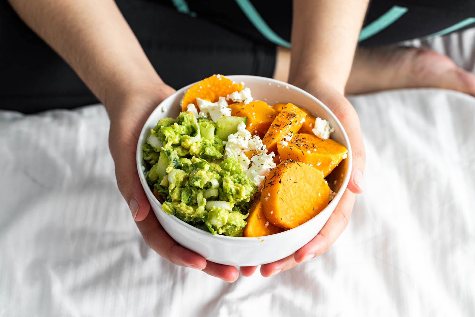 A person holds a bowl containing mashed avocado, sliced sweet potatoes, and crumbled white cheese, sitting on a white bedspread.