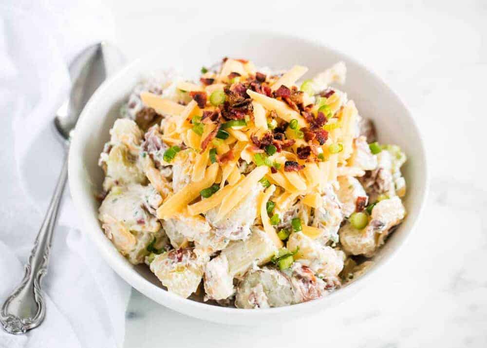 A bowl of creamy potato salad topped with shredded cheese, chopped bacon, and green onions, with a spoon beside the bowl.