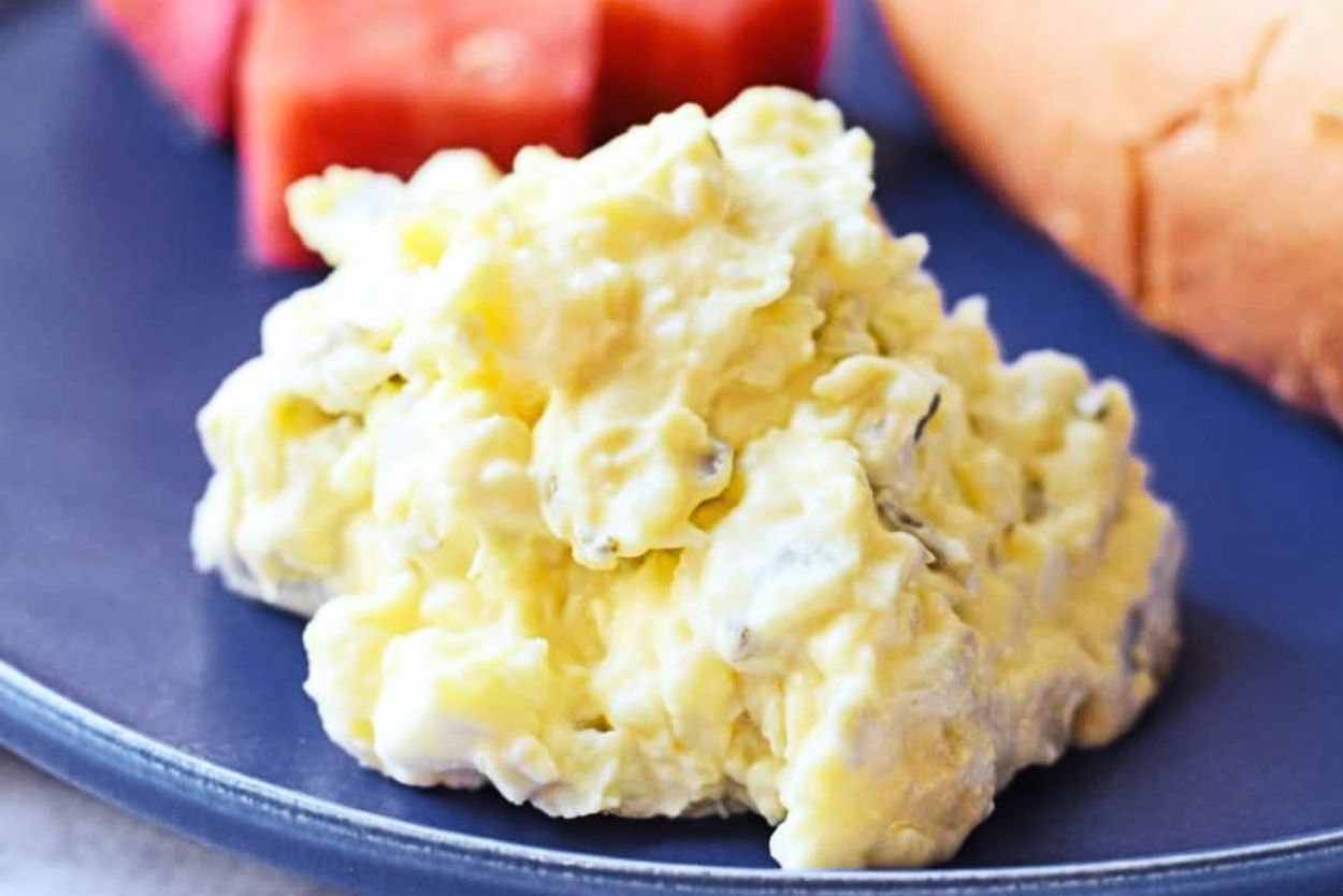 A serving of creamy potato salad on a dark plate, with pieces of watermelon and bread in the background.