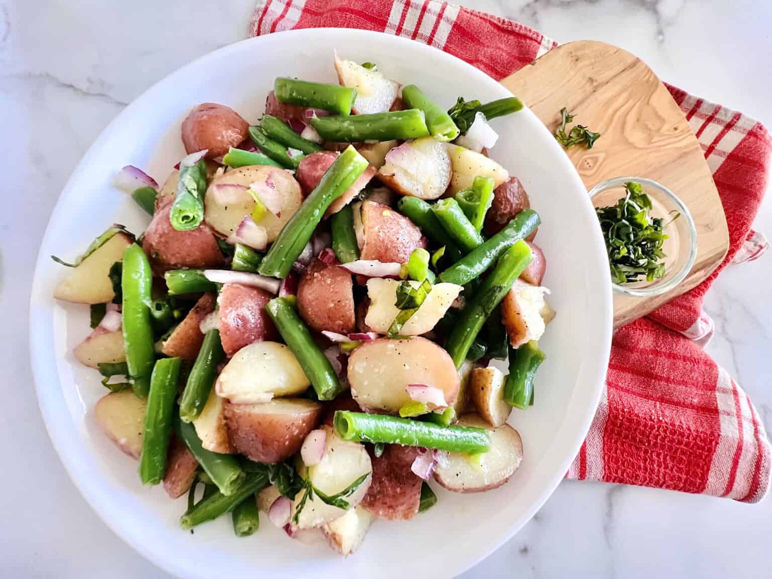 A plate of potato and green bean salad with red onions and herbs sits on a red towel, next to a small dish of chopped herbs on a wooden board.