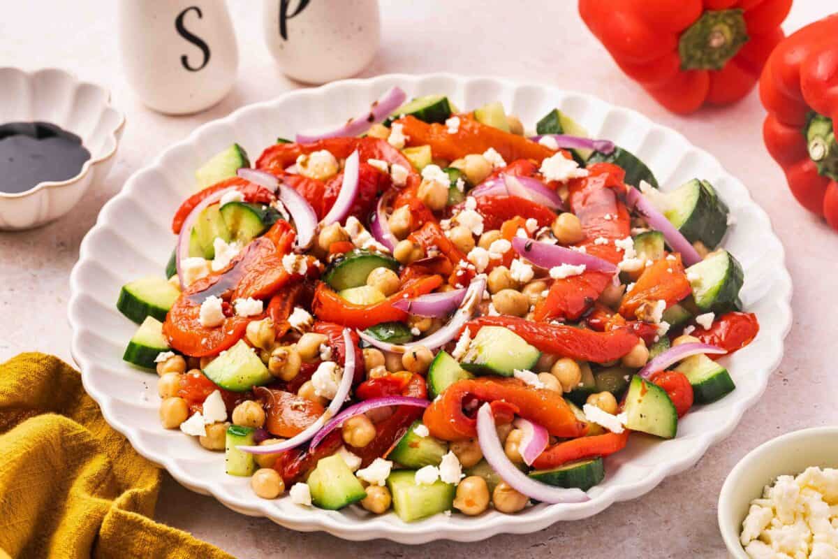 A white plate filled with a colorful salad of chickpeas, cucumbers, roasted red peppers, red onion, and crumbled feta cheese. Salt and pepper shakers and red peppers are in the background.