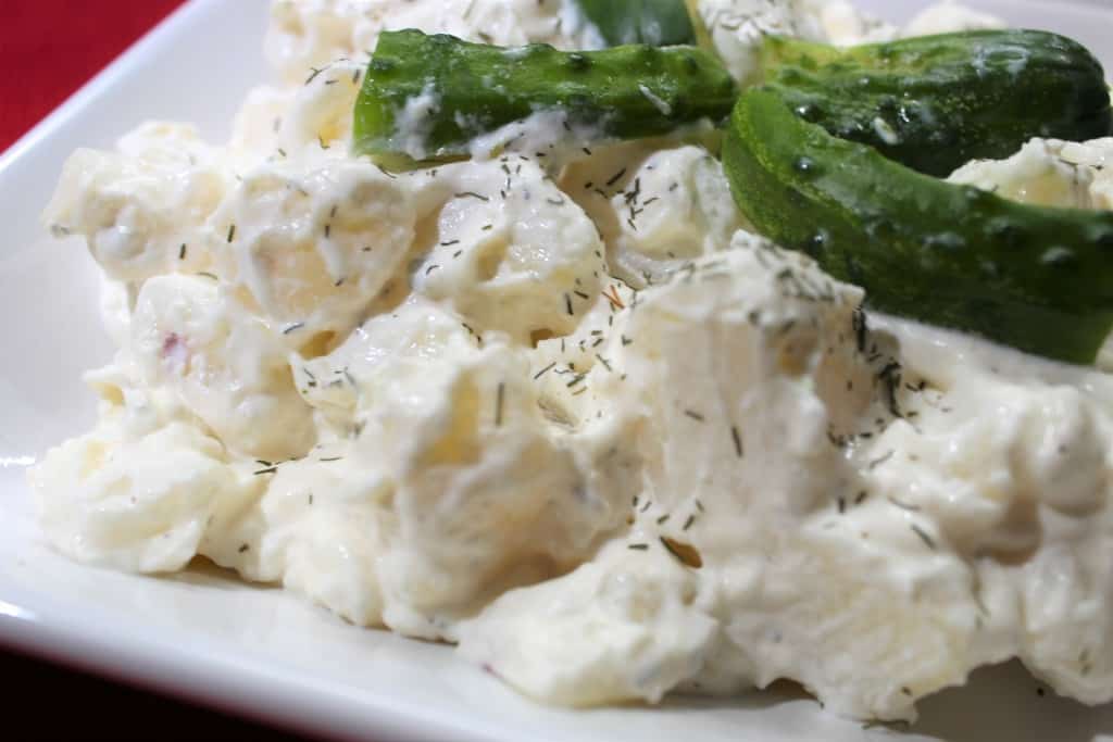 A close-up of creamy potato salad garnished with fresh dill and slices of pickle on a white plate.