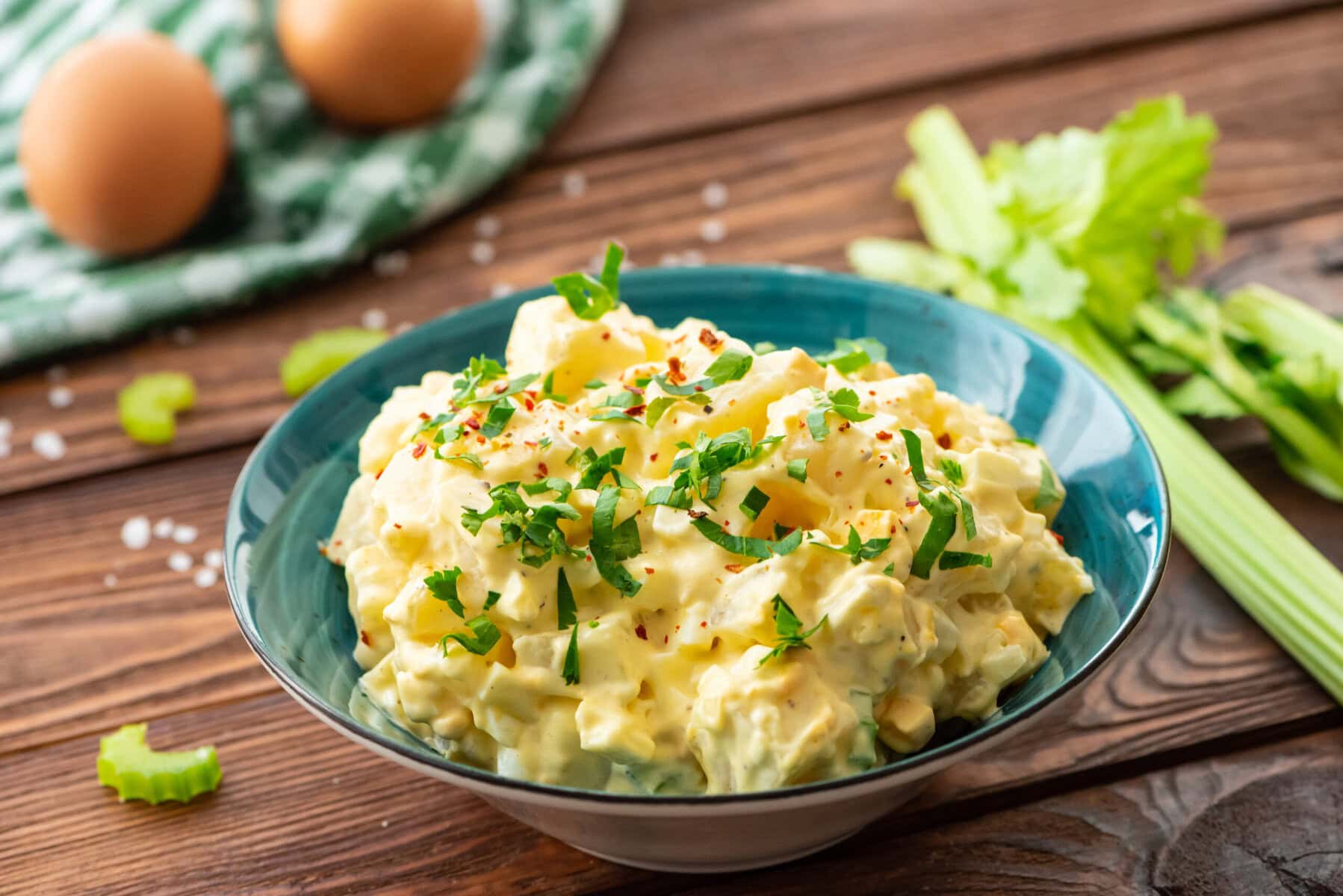 A bowl of creamy egg salad garnished with chopped parsley and paprika sits on a wooden table, with celery stalks and whole eggs in the background.