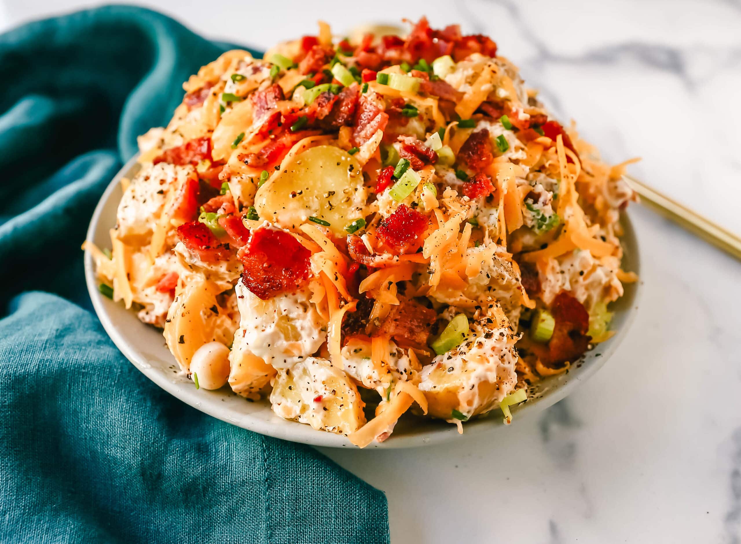A plate of loaded potato salad with chopped bacon, shredded cheese, green onions, and cracked black pepper on a marble surface next to a teal cloth.