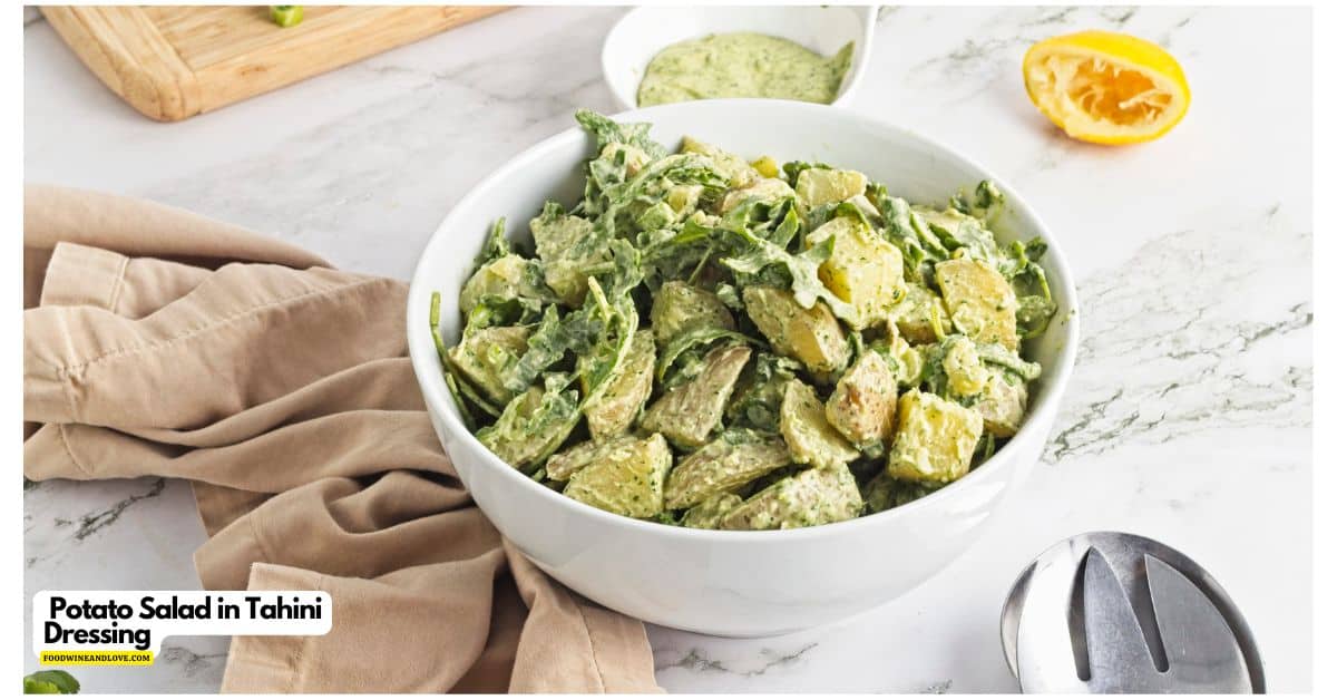 A white bowl filled with potato salad in tahini dressing sits on a marble surface, with a beige napkin, dressing, sliced lemon, and serving utensils nearby.