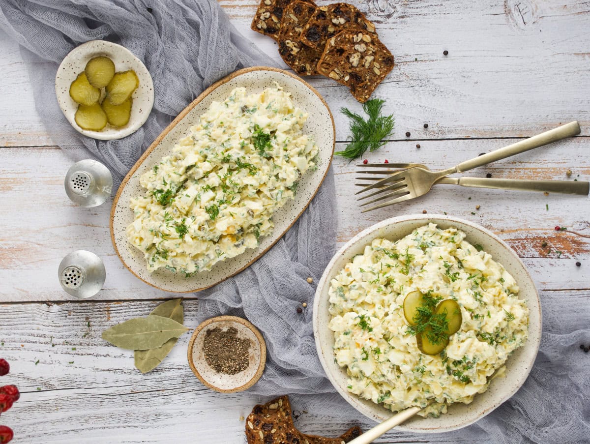 Two plates of creamy egg salad topped with herbs and pickle slices, served with crispbread, pepper, and sliced pickles on a rustic white wooden table.