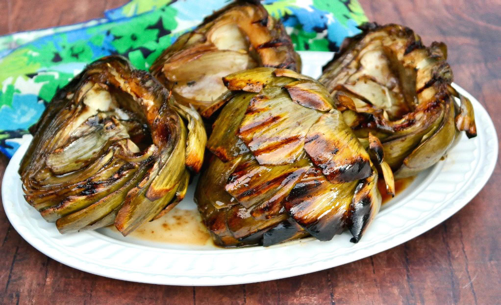 Four grilled artichoke halves are arranged on a white oval plate, placed on a wooden surface with a blue and green floral cloth in the background.