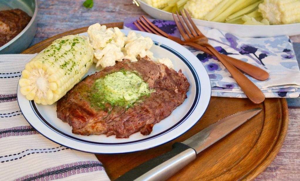 A plate with grilled steak topped with herb butter, corn on the cob, and macaroni salad sits on a wooden tray with a knife and copper utensils next to it.