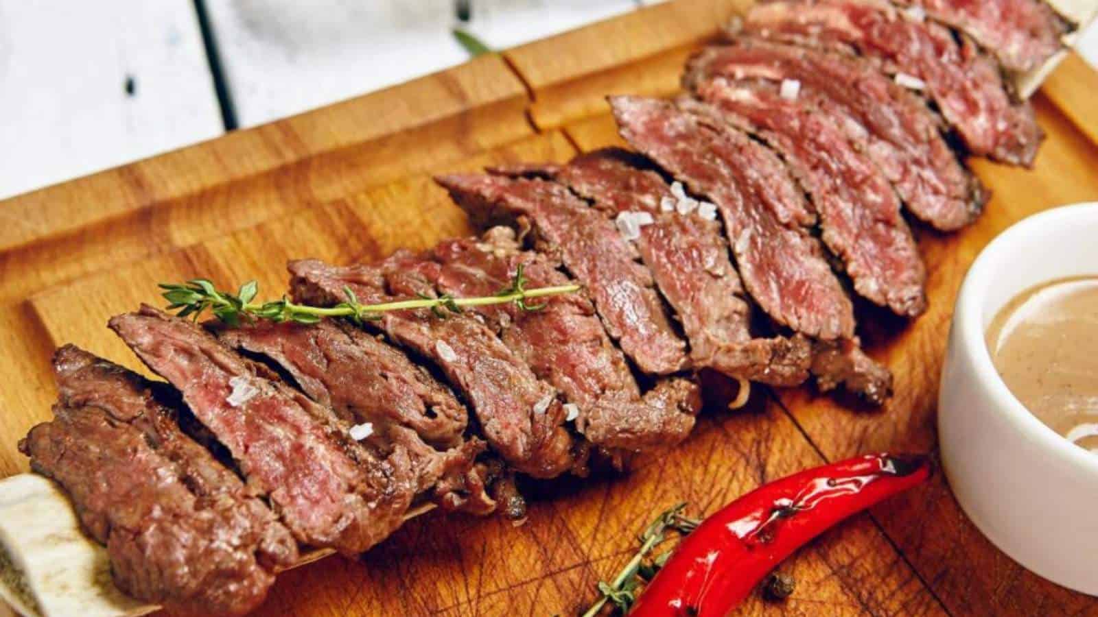 Sliced grilled steak served on a wooden board with coarse salt, fresh herbs, a red chili pepper, and a cup of dipping sauce.