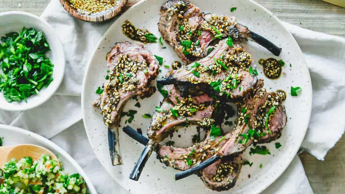 A white plate with grilled lamb chops topped with sesame seeds and chopped herbs, surrounded by small bowls of herbs and salad.