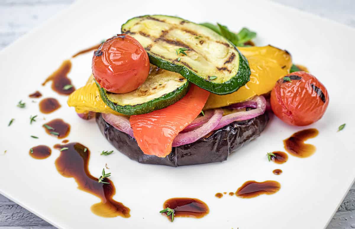 A stack of grilled vegetables, including zucchini, tomato, bell pepper, and eggplant, served on a white plate with balsamic glaze drizzled around.