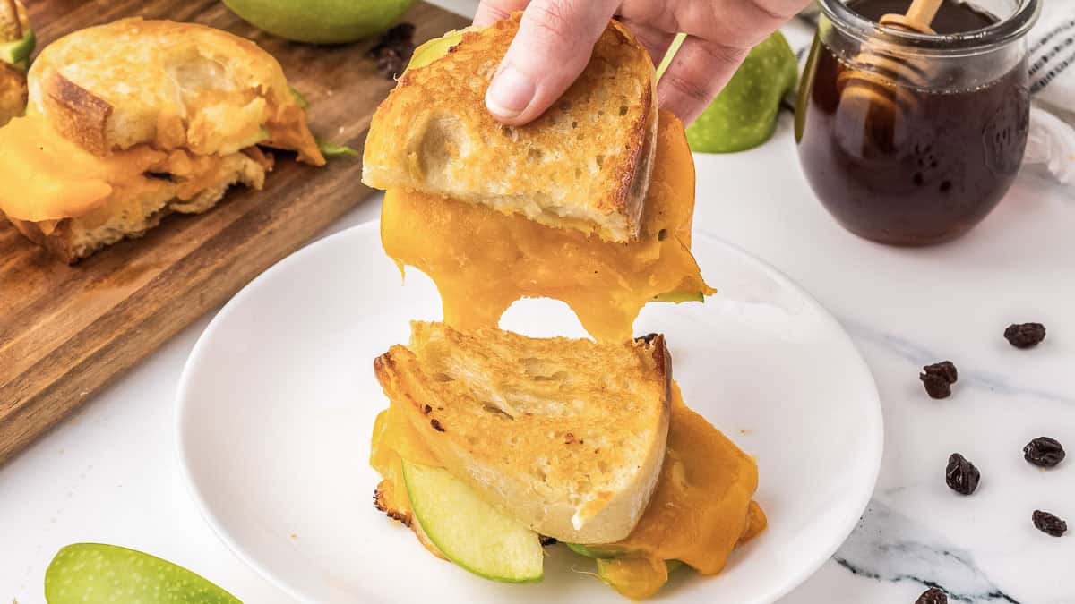 A hand pulls apart a grilled cheese sandwich with melted cheddar and green apple slices on a white plate; apple slices and a jar of honey are nearby.