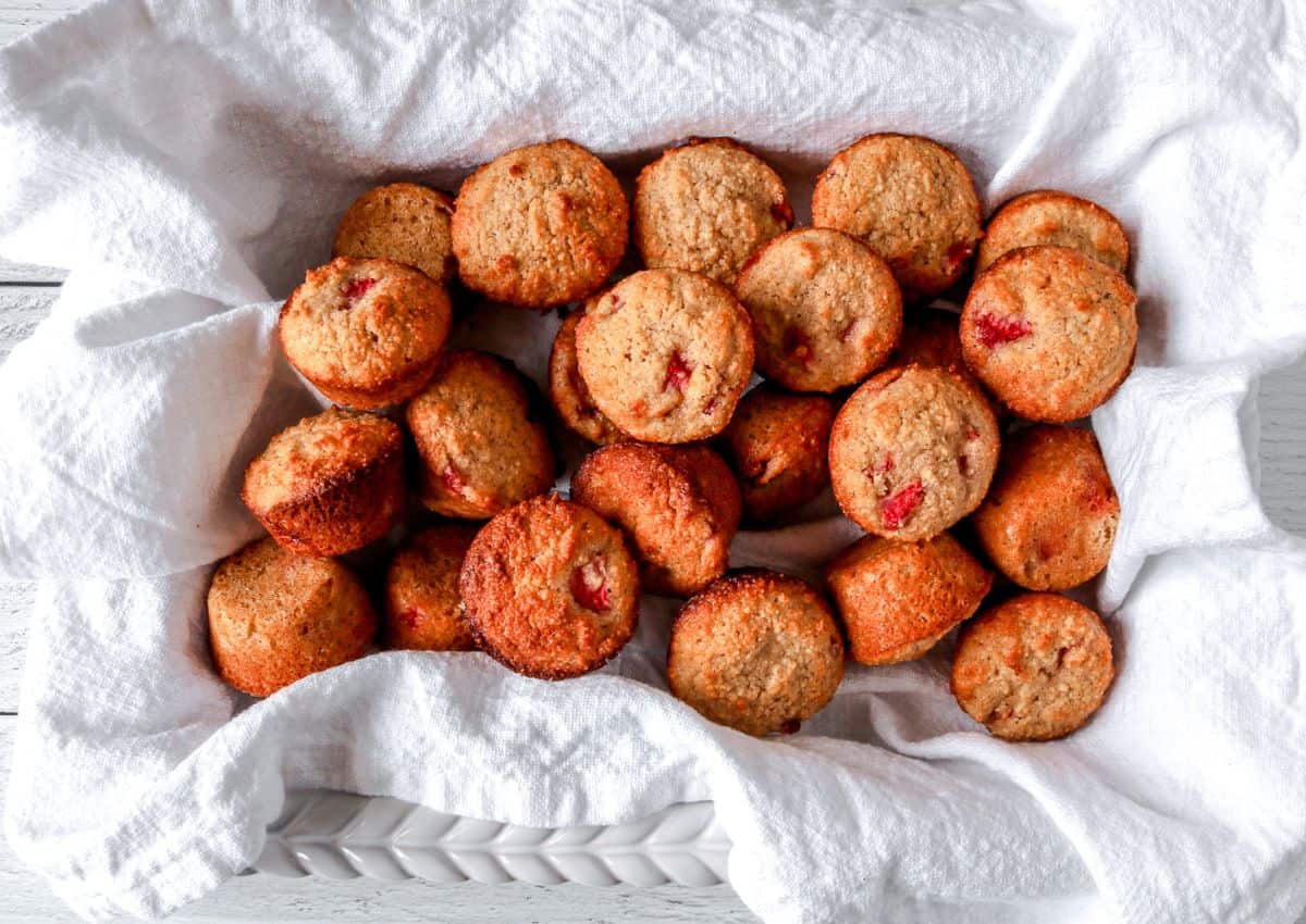A basket lined with a white cloth holds several golden-brown muffins with visible pieces of fruit.