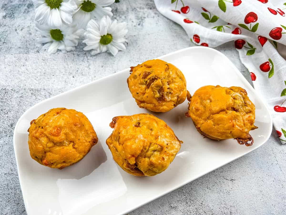 Four savory muffins are arranged on a white rectangular plate, with a floral napkin and white flowers in the background.