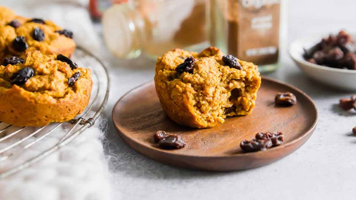 A pumpkin muffin with raisins sits on a wooden plate, partially bitten, with more muffins on a cooling rack and a bowl of raisins in the background.