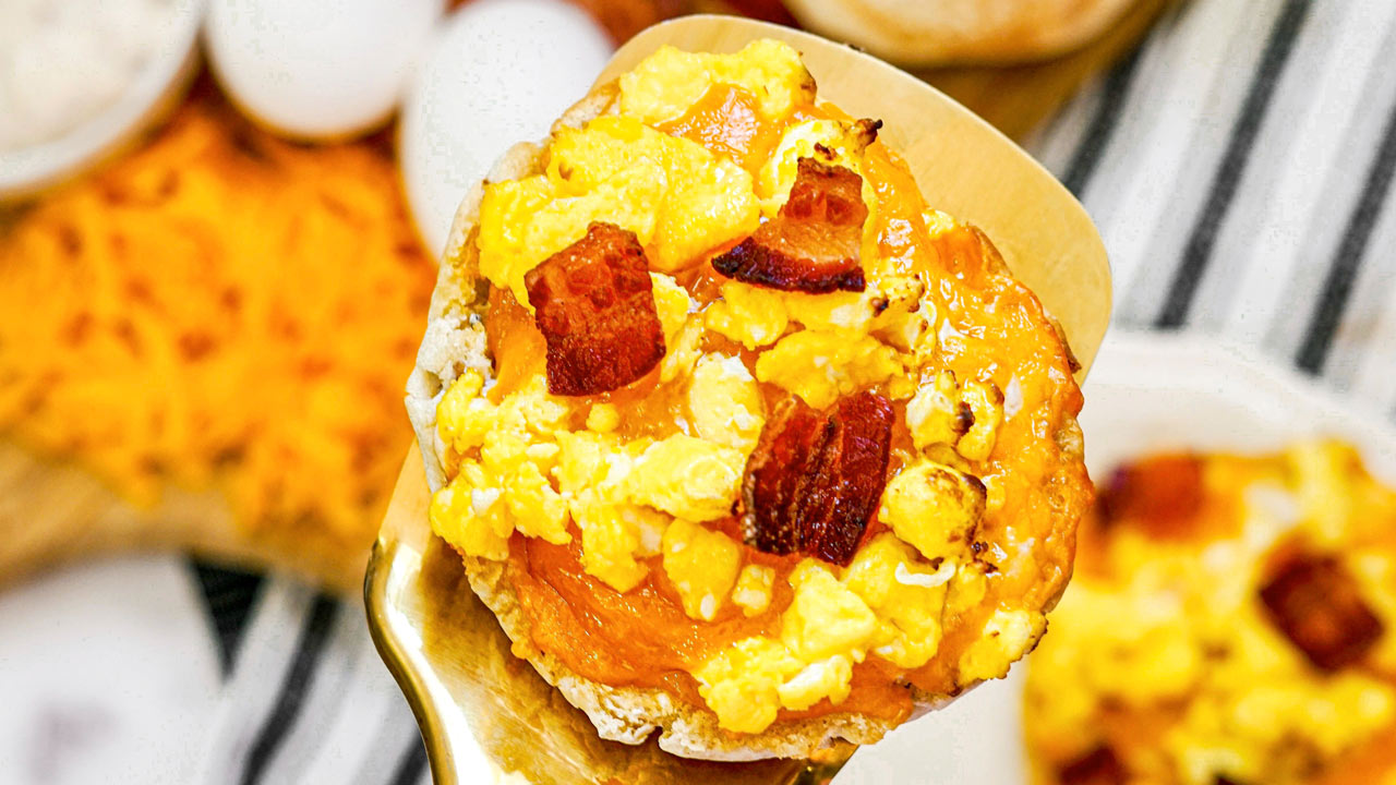 A close-up of a breakfast muffin topped with scrambled eggs, melted cheddar cheese, and bacon pieces on a spatula.