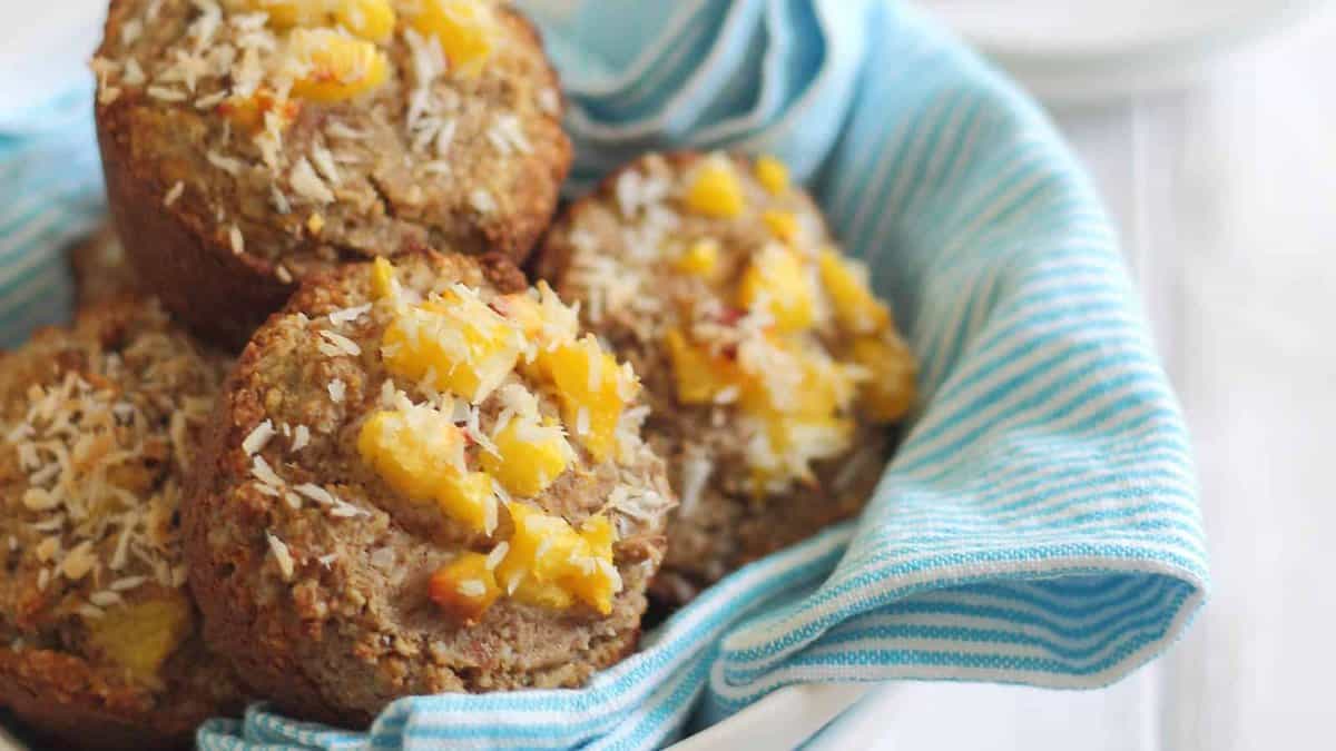 A close-up of several muffins topped with chopped fruit and shredded coconut, arranged in a bowl lined with a blue and white striped cloth.