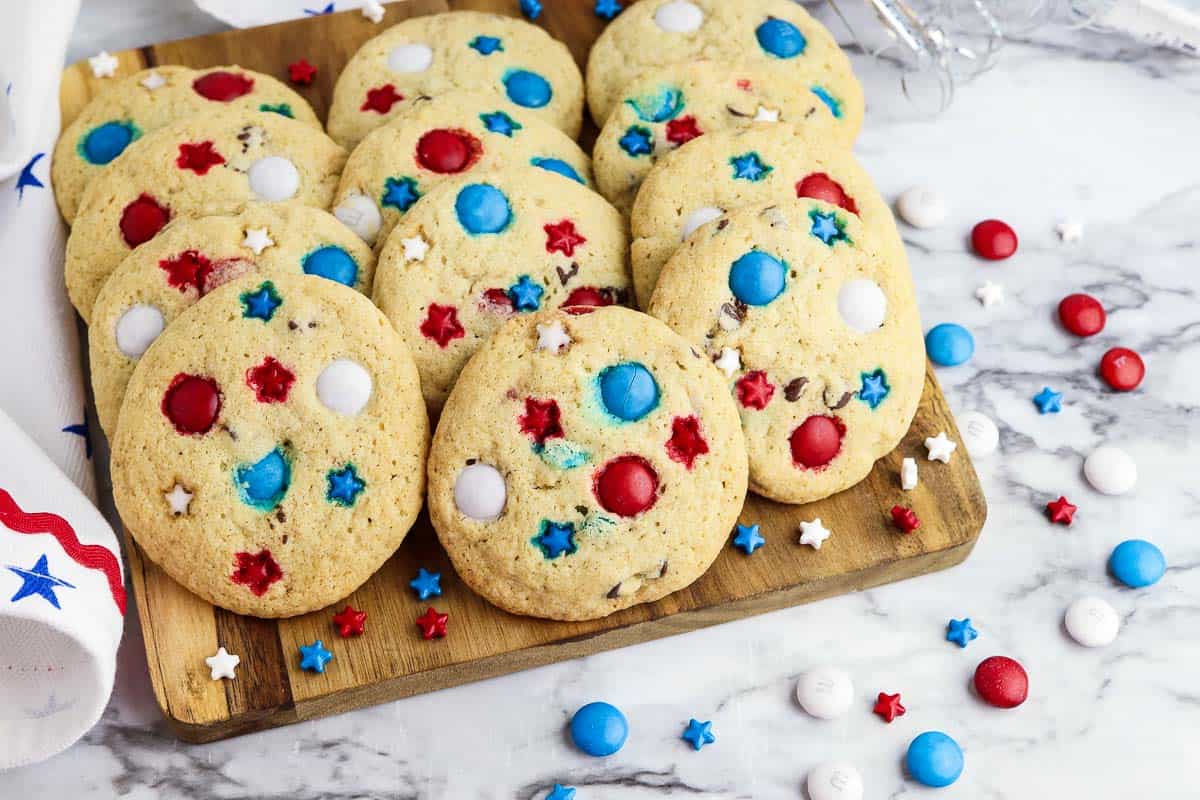 A wooden board holds cookies topped with red, white, and blue candies and star-shaped sprinkles; more candies are scattered on a marble surface nearby.