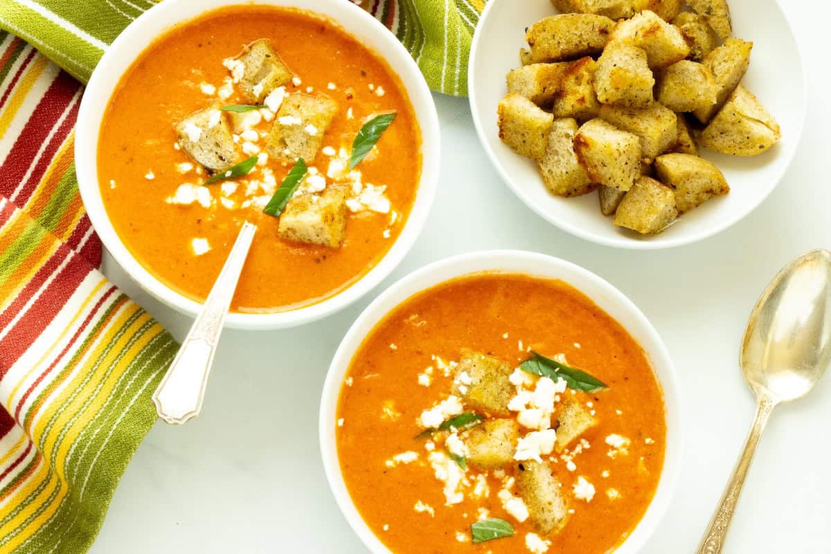 Two bowls of tomato soup topped with croutons, cheese, and basil, with an additional bowl of croutons and a striped napkin beside them.