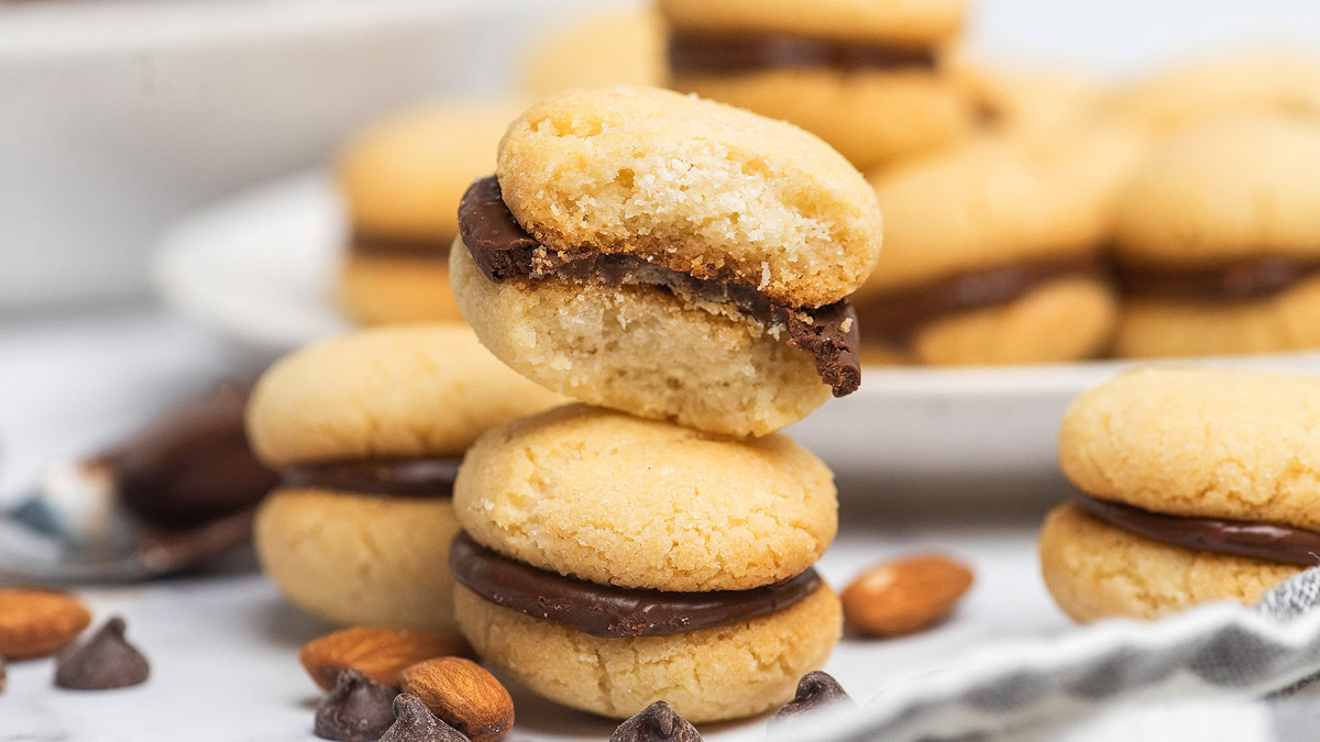 Two sandwich cookies with chocolate filling are stacked on top of each other, with whole almonds and chocolate chips scattered around and more cookies in the background.