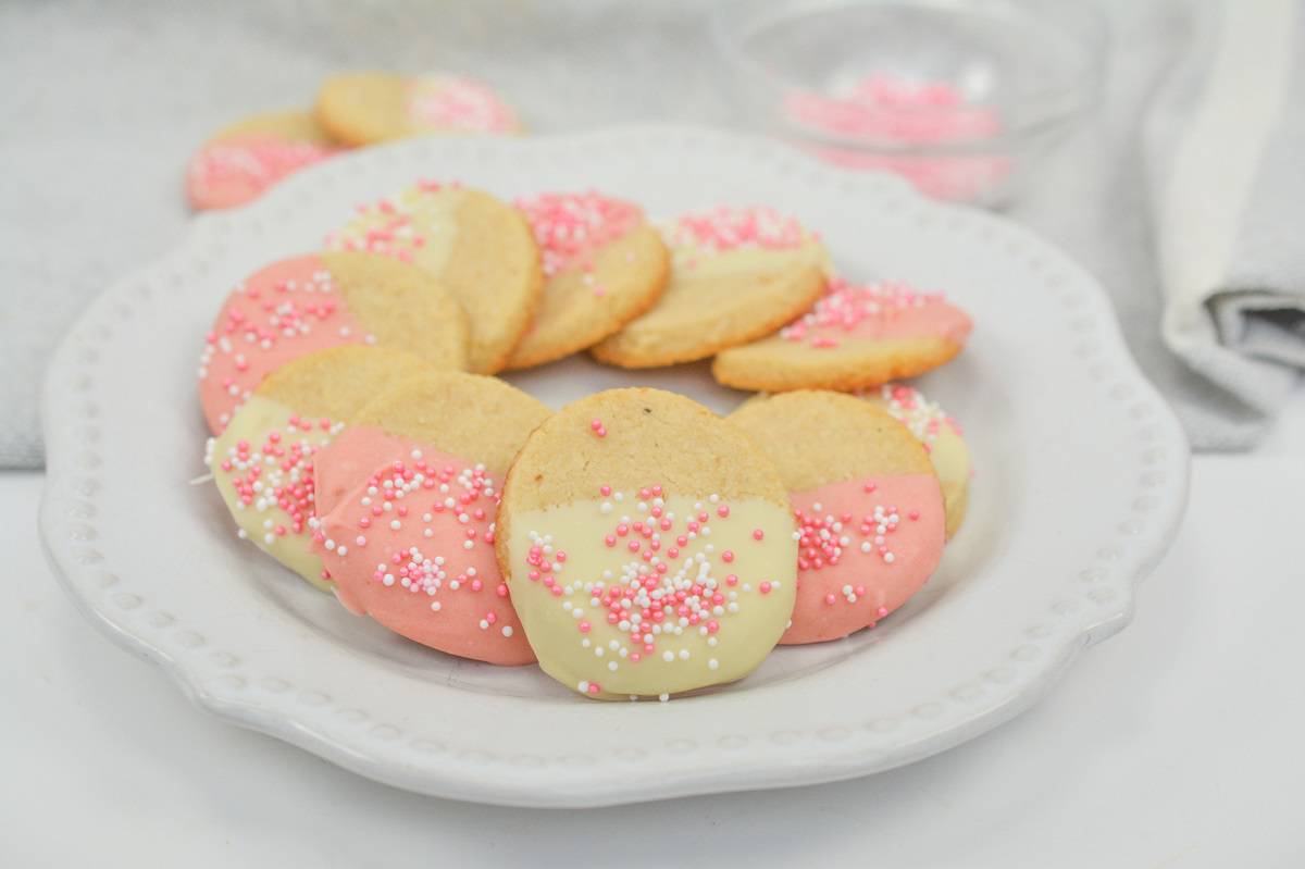 A white plate with cookies dipped in pink and white icing, topped with pink and white sprinkles, arranged in a circle.