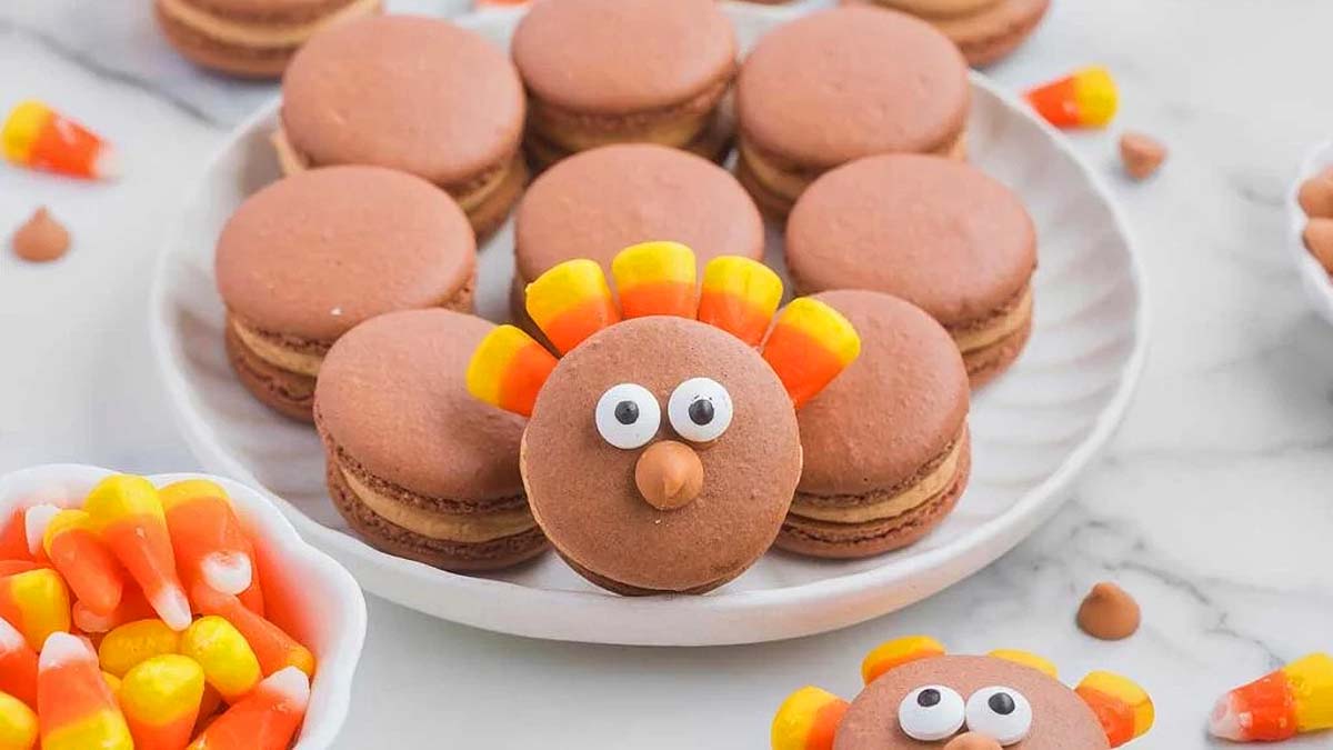 Chocolate macarons are arranged on a plate, with one decorated as a turkey using candy eyes, candy corn, and a brown candy nose. Bowls of candy corn are nearby.