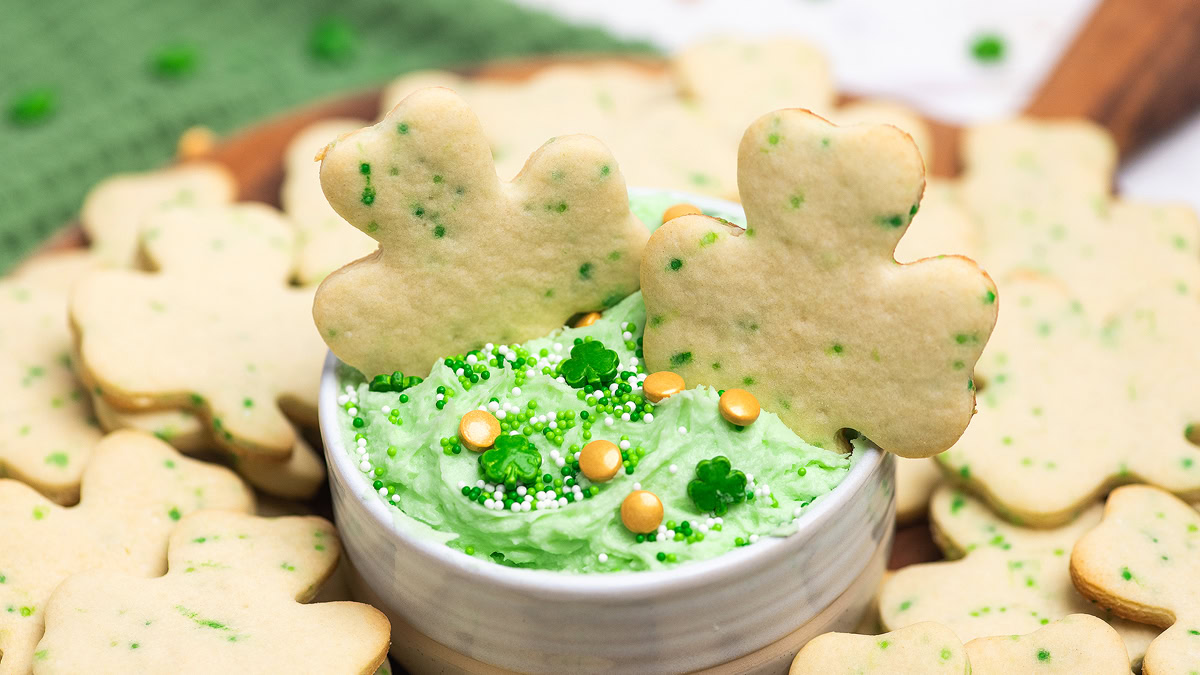 Shamrock-shaped cookies with green sprinkles are arranged around and in a bowl of green-frosted dip, garnished with gold and white sprinkles.