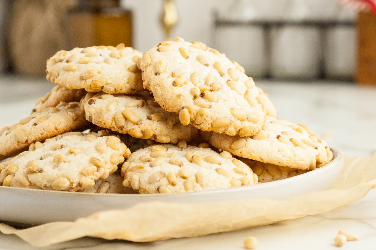 A plate of cookies topped with pine nuts is placed on a piece of parchment paper on a light-colored surface.