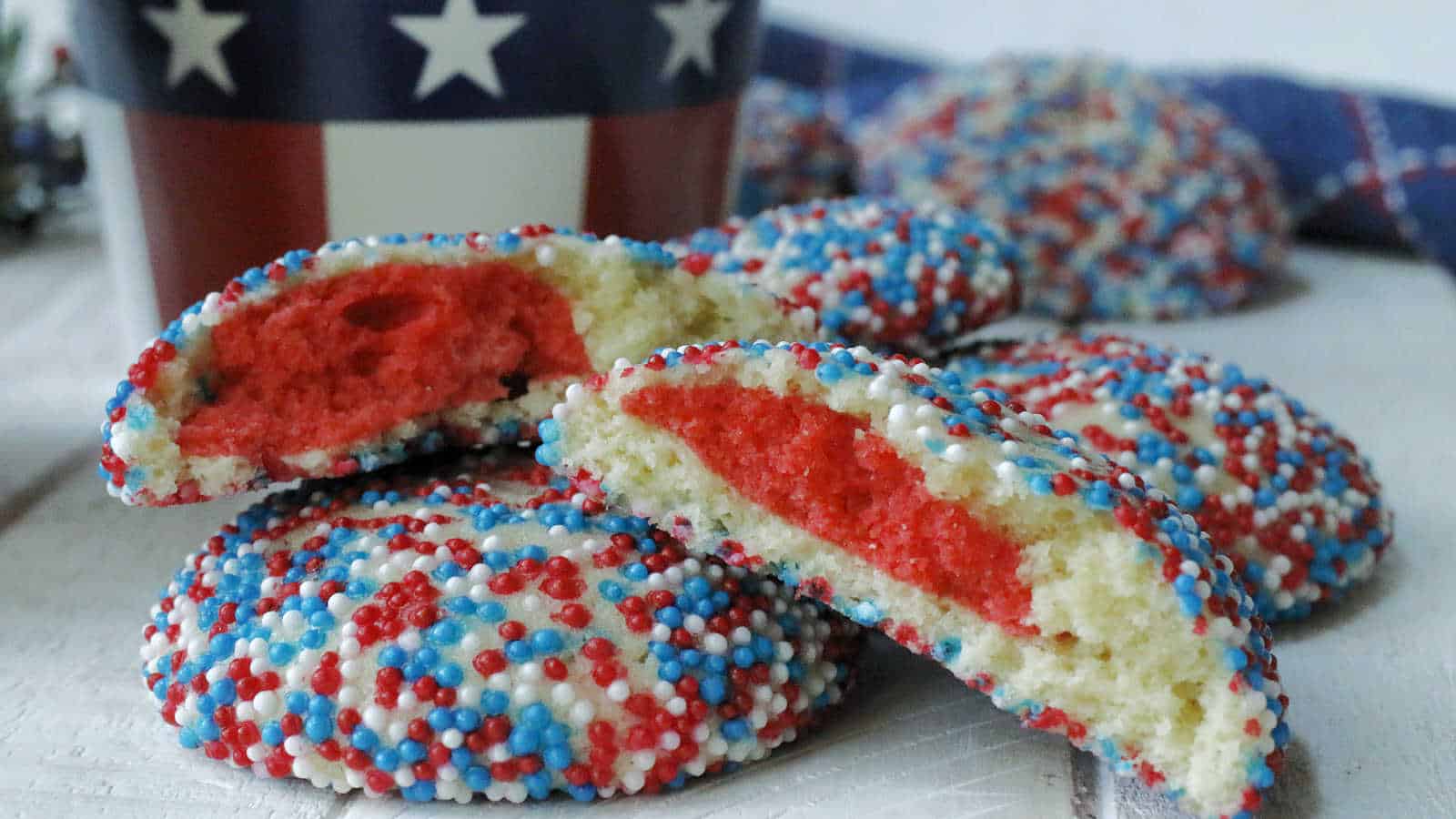Close-up of cookies coated in red, white, and blue sprinkles with a red center, arranged on a white surface. A patriotic cup with stars is visible in the background.