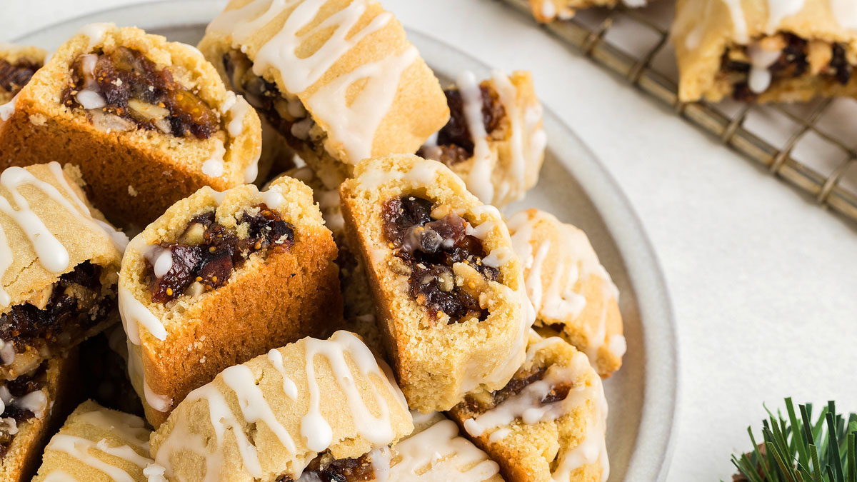 A plate of rectangular pastries filled with a dark fruit and nut mixture, drizzled with white icing, sits on a light surface next to a cooling rack with more pastries.