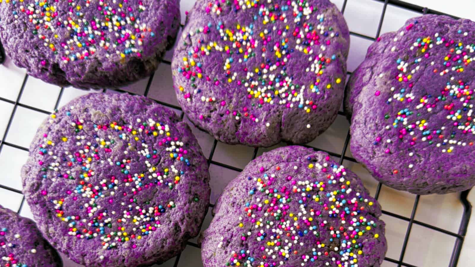 Purple cookies with colorful sprinkles are cooling on a black wire rack.