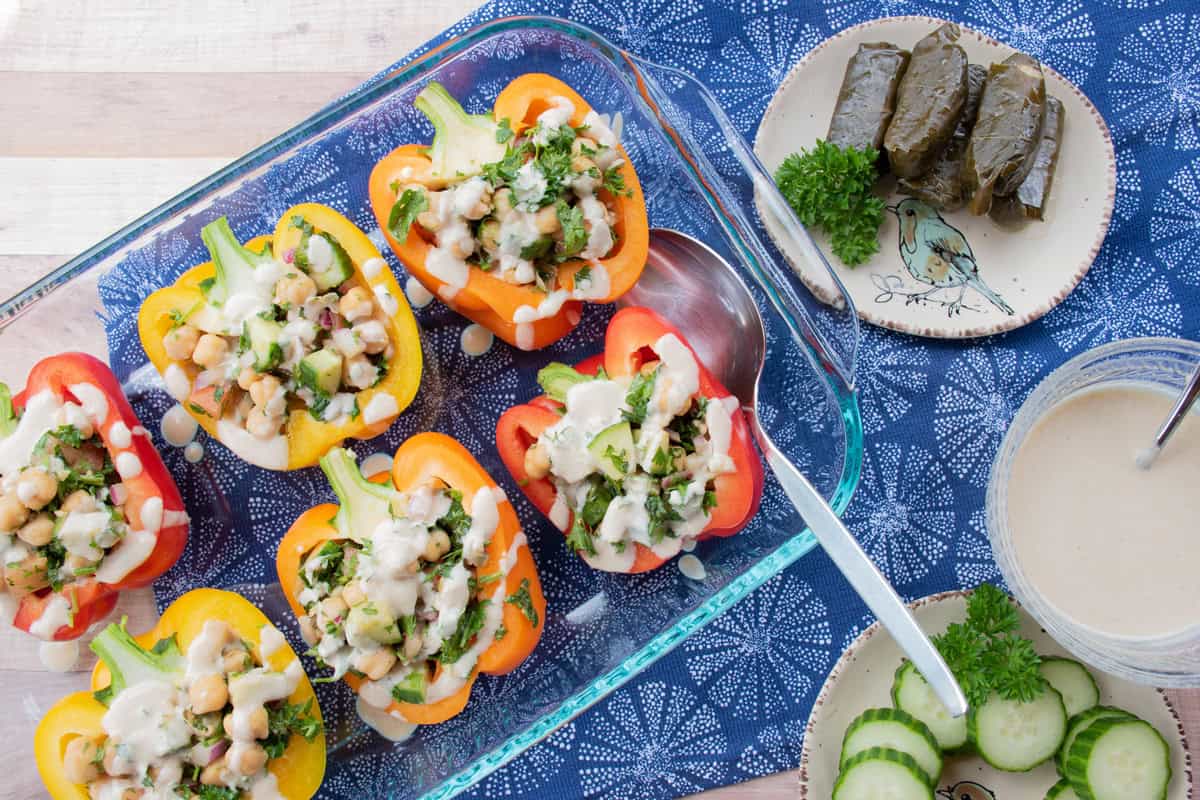 Halved bell peppers stuffed with chickpeas and vegetables, drizzled with sauce in a glass dish, served alongside grape leaves, cucumber slices, and parsley on a blue tablecloth.
