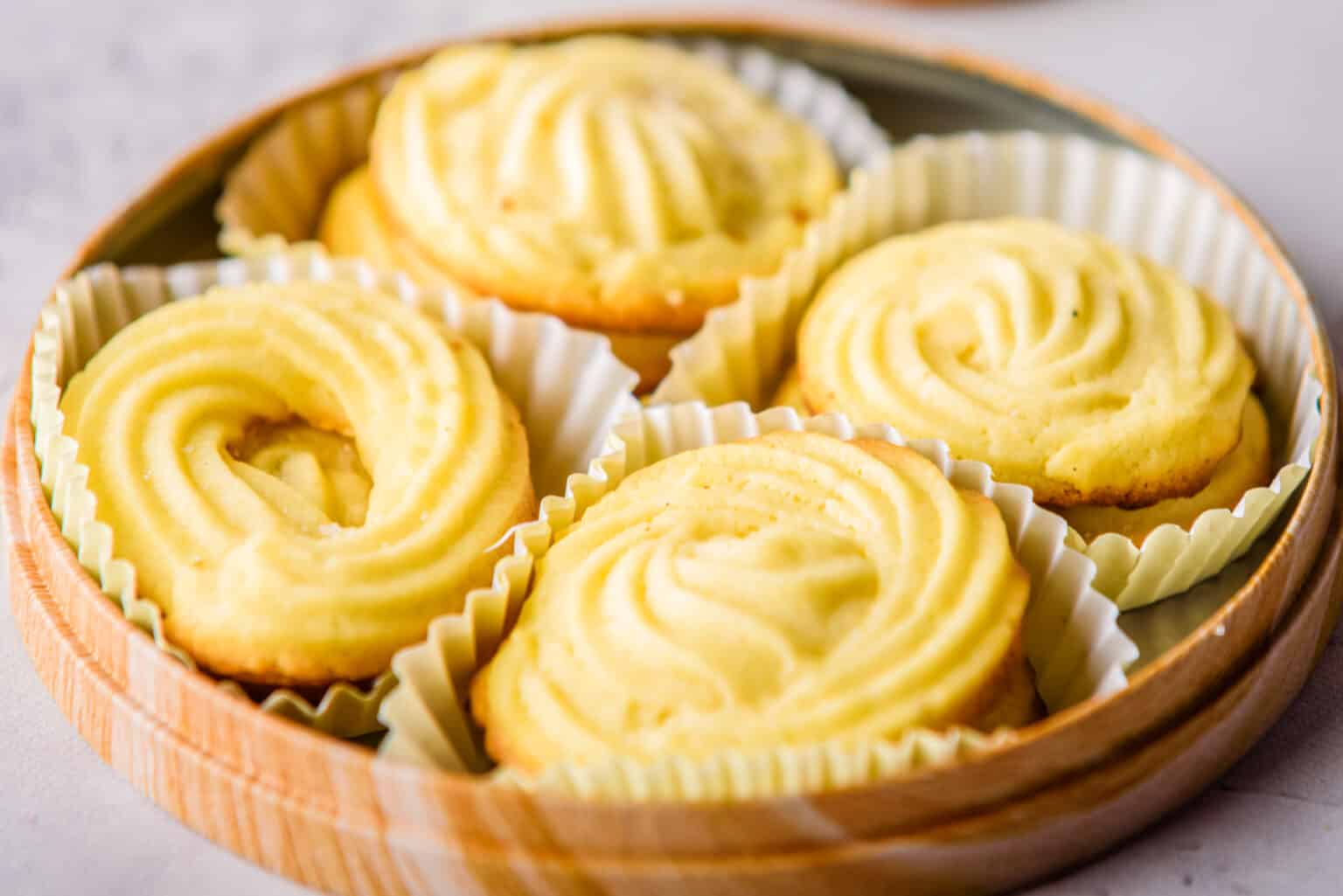 Four butter cookies with a swirled pattern are placed in white paper liners inside a round wooden container.