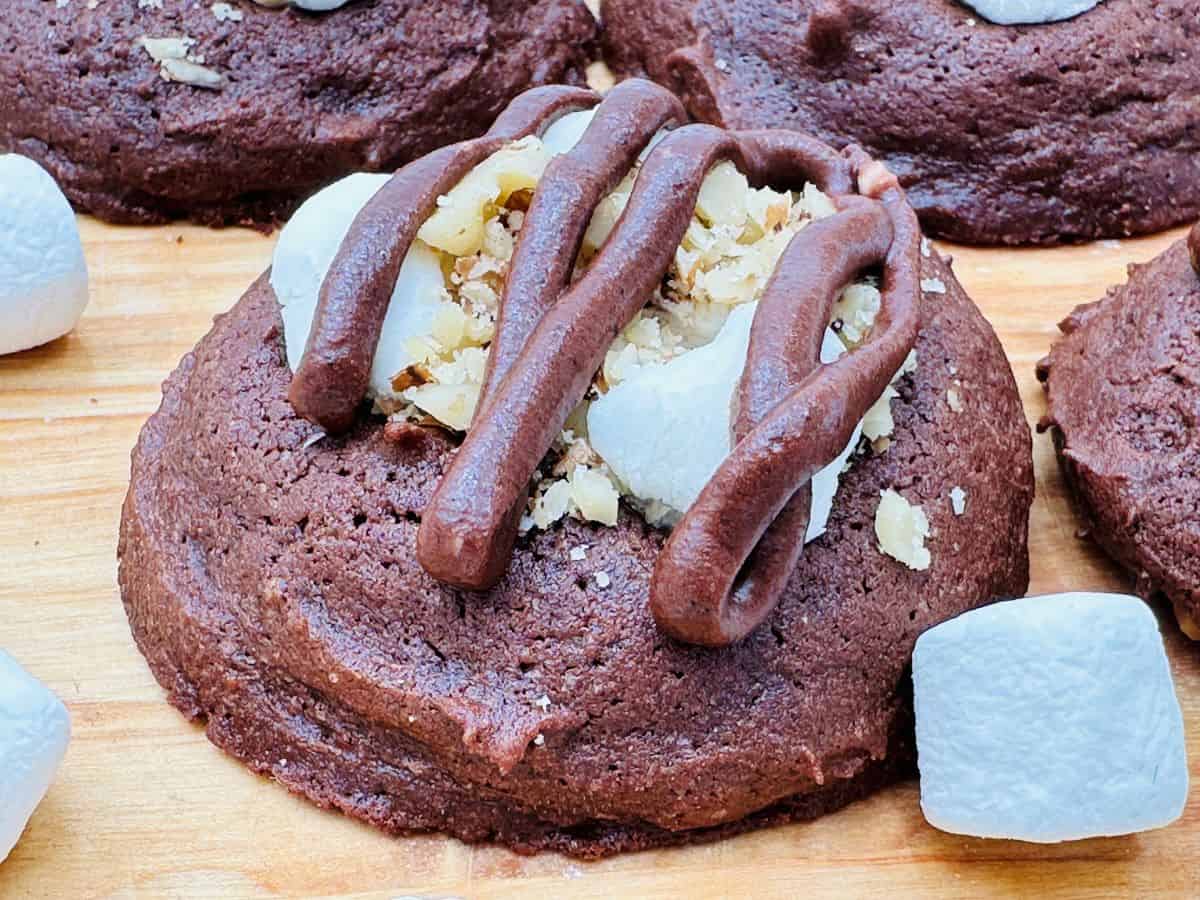 A close-up of a chocolate cookie topped with chopped nuts, marshmallows, and chocolate drizzle, placed on a wooden surface with marshmallows nearby.
