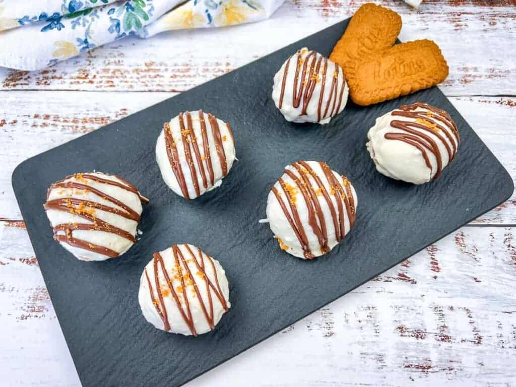Six round, white chocolate-coated desserts drizzled with milk chocolate, arranged on a black slate tray with two rectangular cookies in the corner.
