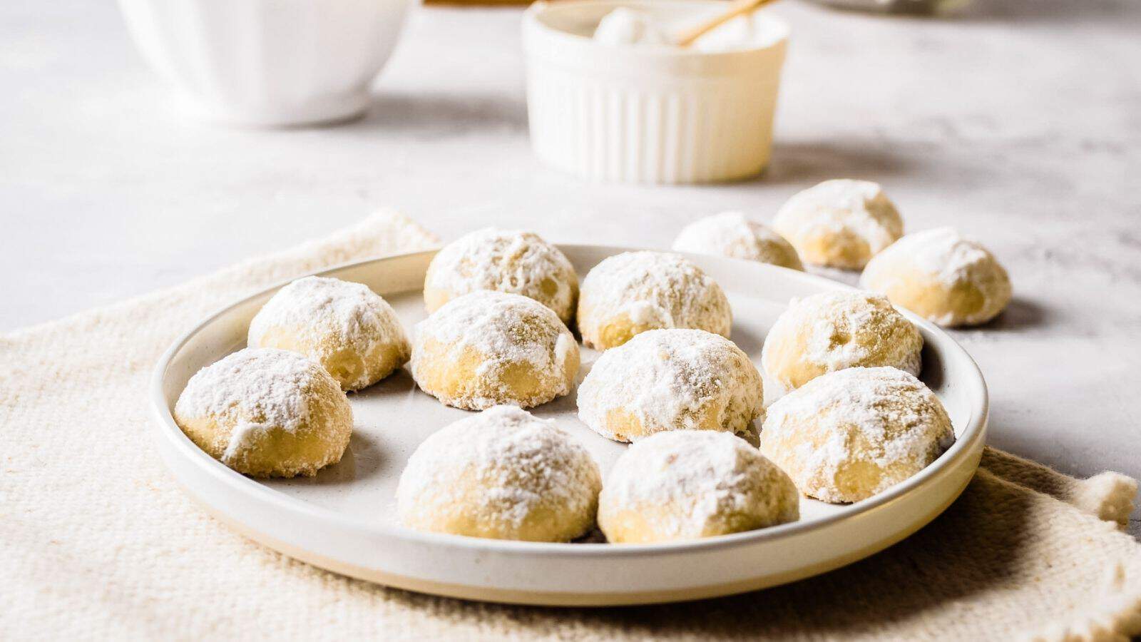 A plate of round, powdered sugar-covered cookies sits on a light-colored surface with more cookies and a ramekin in the background.