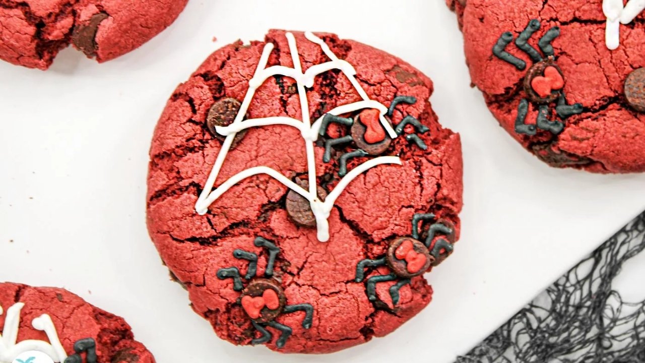 A red, cracked cookie decorated with white icing in a web pattern and black and red icing shaped like spiders on a white surface.