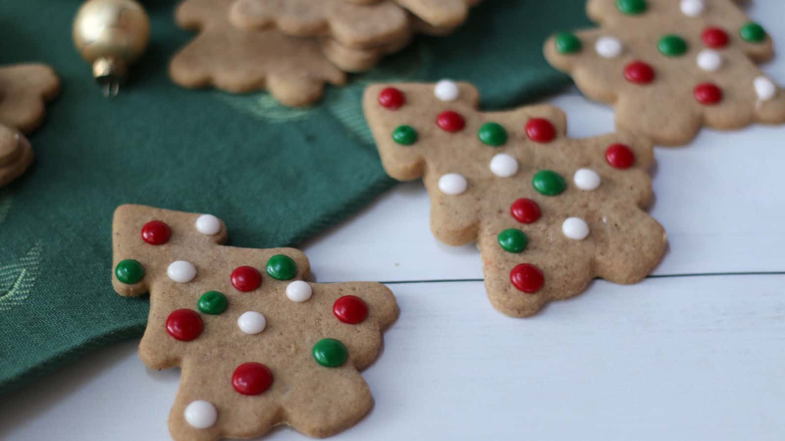 Three Christmas tree-shaped cookies decorated with red, white, and green candy dots are placed on a white surface next to a green cloth.