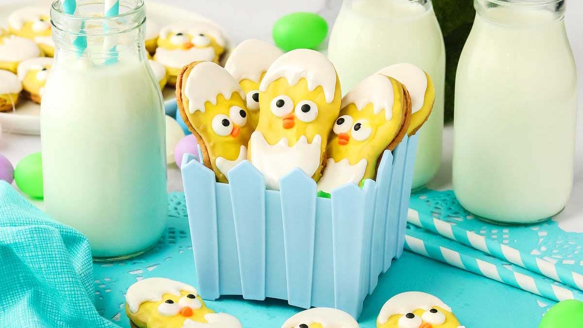 Decorated cookies shaped like chicks in eggshells are arranged in a blue container, with bottles of milk and pastel decorations around them.