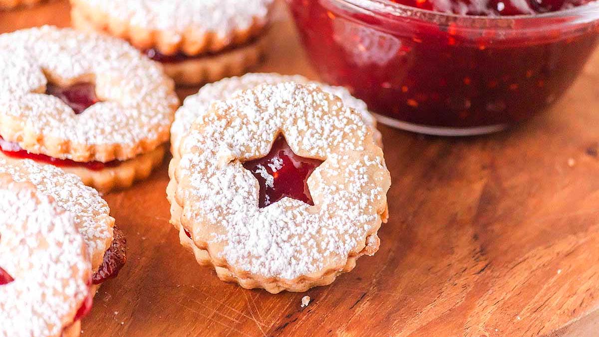 Round sandwich cookies dusted with powdered sugar and filled with red jam, with a star-shaped cutout in the center, are arranged on a wooden surface next to a bowl of jam.