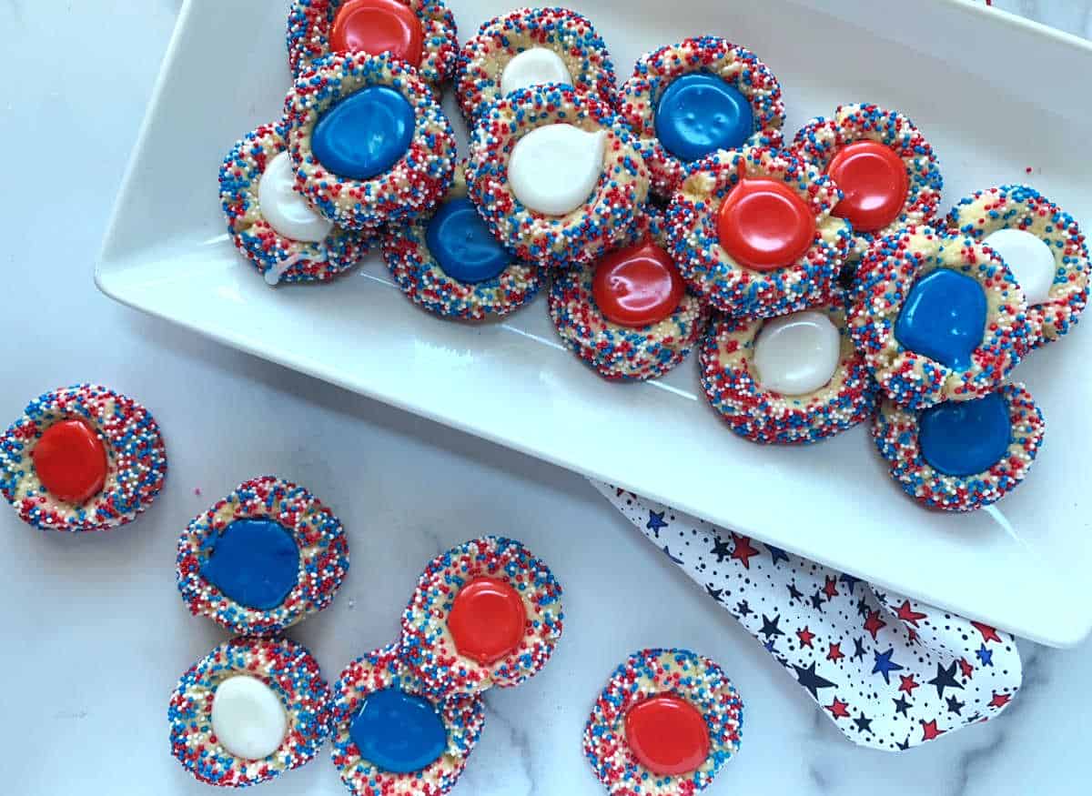 A plate of thumbprint cookies decorated with red, white, and blue sprinkles and filled with red, white, or blue centers, arranged on a white surface.
