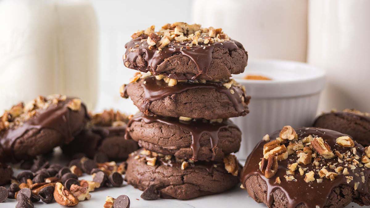 A stack of chocolate cookies topped with chopped nuts and drizzled chocolate, surrounded by chocolate chips and milk in the background.