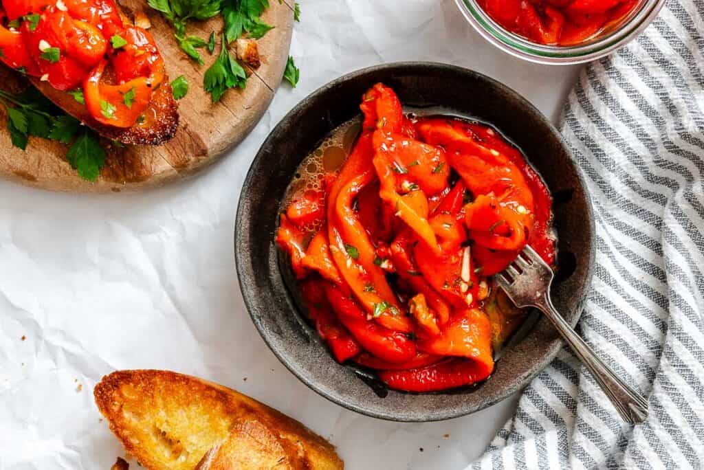 A bowl of roasted red peppers in oil with herbs, next to toasted bread slices and a wooden board with bruschetta.