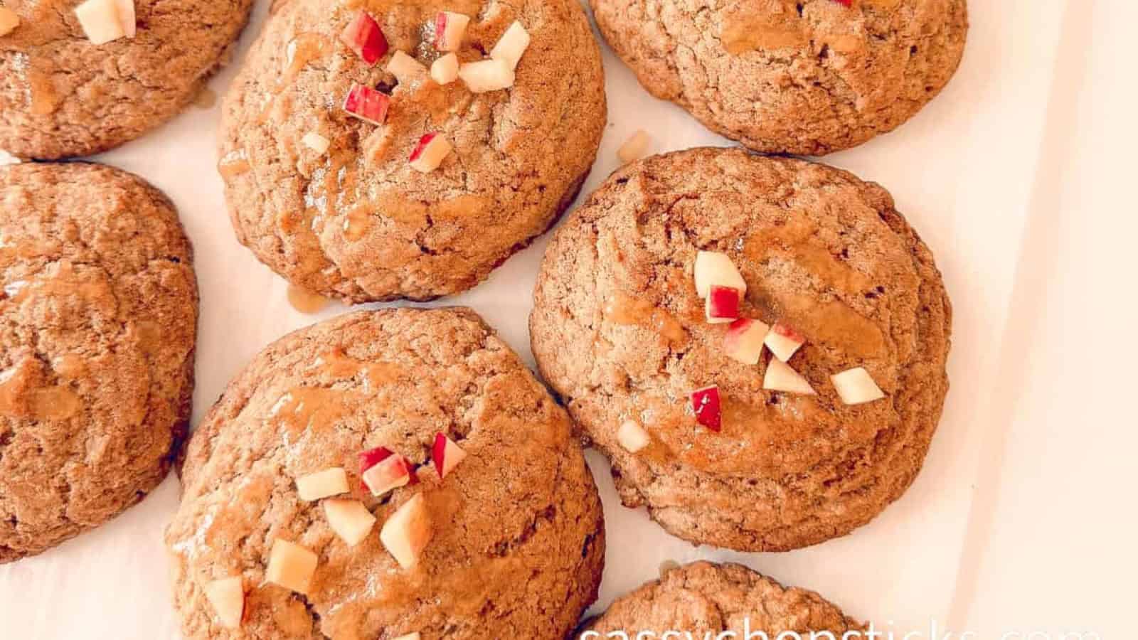 Seven large cookies topped with small diced red and white apple pieces are arranged on a white surface.