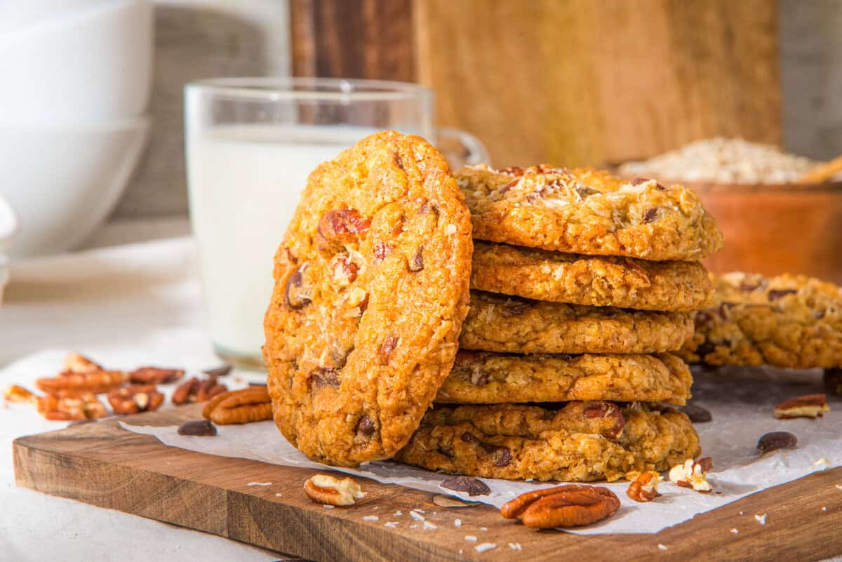 A stack of oatmeal cookies with pecans and chocolate chips on a wooden board, with a glass of milk and scattered pecans nearby.
